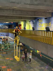 Un niño monta una bicicleta amarilla suspendida en una exposición de cable alto sobre una pasarela del museo, mientras los visitantes caminan por debajo a través de una zona con red. Luces de colores y una exposición mural con temática científica titulada «¡Carga tus pilas!» decoran el espacio.