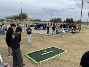 Evento al aire libre en un parque con grupos de personas reunidas alrededor de mesas y estaciones de actividades cerca de una cancha de baloncesto. En primer plano, dos personas están de pie cerca de una pequeña pista de carreras con bordes de espuma sobre el césped, con coches de juguete colocados en su interior. Una persona en segundo plano está tomando una foto, y se ven tiendas de campaña, conos y diversas instalaciones para actividades en toda la zona bajo un cielo nublado.
