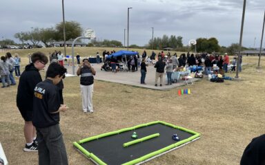 Evento al aire libre en un parque con grupos de personas reunidas alrededor de mesas y estaciones de actividades cerca de una cancha de baloncesto. En primer plano, dos personas están de pie cerca de una pequeña pista de carreras con bordes de espuma sobre el césped, con coches de juguete colocados en su interior. Una persona en segundo plano está tomando una foto, y se ven tiendas de campaña, conos y diversas instalaciones para actividades en toda la zona bajo un cielo nublado.